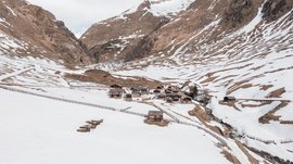 Malga Fane, il più bel villaggio alpino dell’Alto Adige L'immagine mostra un piccolo insediamento di case tradizionali in legno situato in una valle, circondato da montagne innevate. La neve inizia a sciogliersi e emergono delle superfici montuose marroni, mentre una recinzione delimita l'area.