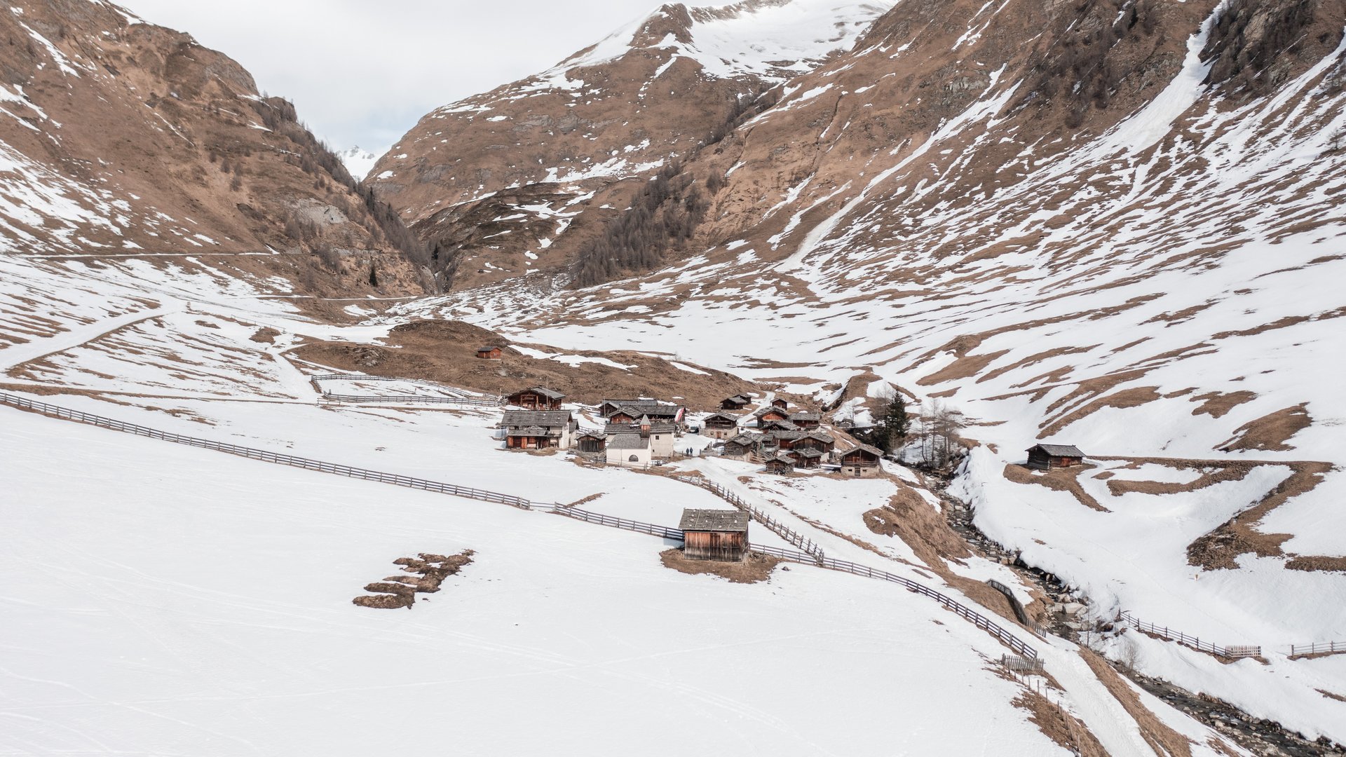 Malga Fane, il più bel villaggio alpino dell’Alto Adige L'immagine mostra un piccolo insediamento di case tradizionali in legno situato in una valle, circondato da montagne innevate. La neve inizia a sciogliersi e emergono delle superfici montuose marroni, mentre una recinzione delimita l'area.