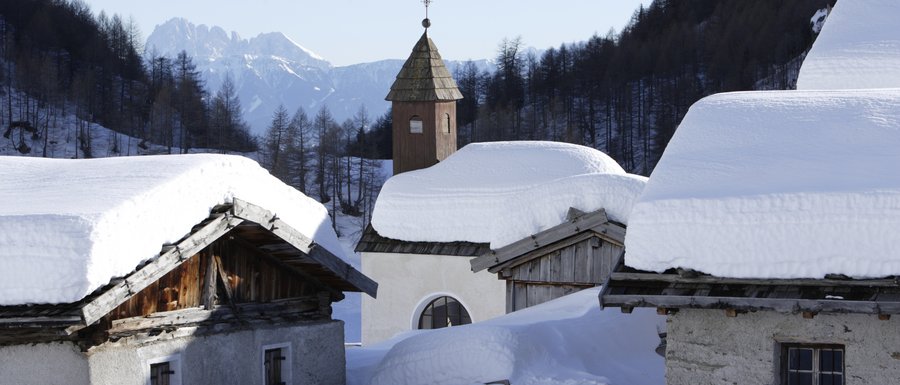 Benvenuti nell’Area Vacanze Sci e Malghe Rio Pusteria! L'immagine mostra un villaggio di baite di montagna innevato con neve profonda sui tetti degli edifici. Sullo sfondo si vede una piccola torre campanaria con tetto a punta, circondata da alberi e montagne coperti di neve.