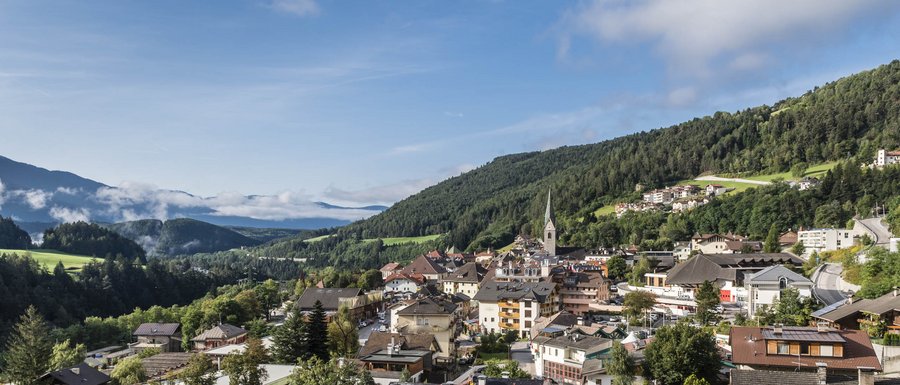 Benvenuti nell’Area Vacanze Sci e Malghe Rio Pusteria! L'immagine mostra una piccola città pittoresca in una valle verde, circondata da colline boscose e montagne. Al centro dell'immagine si erge un campanile con una cuspide appuntita, e gli edifici sono circondati da una vegetazione rigogliosa e alcune nuvole sparse.