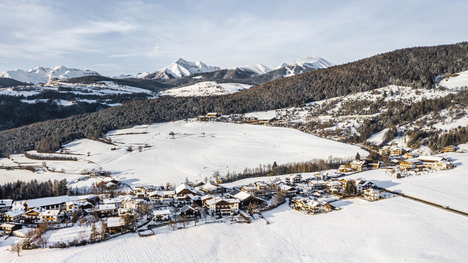 Il paradiso escursionistico di Rodengo L'immagine mostra un paesaggio innevato con un villaggio che si estende in primo piano. Sullo sfondo si vedono colline boschive e montagne innevate sotto un cielo limpido. Le case del villaggio sono parzialmente coperte di neve, e la scena appare tranquilla e pacifica.