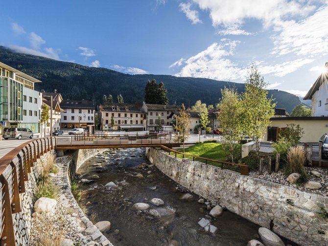 Il paradiso escursionistico di Rodengo L'immagine mostra un piccolo ponte che attraversa un ruscello limpido in un villaggio. Sullo sfondo si vedono case e colline o montagne verdi sotto un cielo blu con poche nuvole.