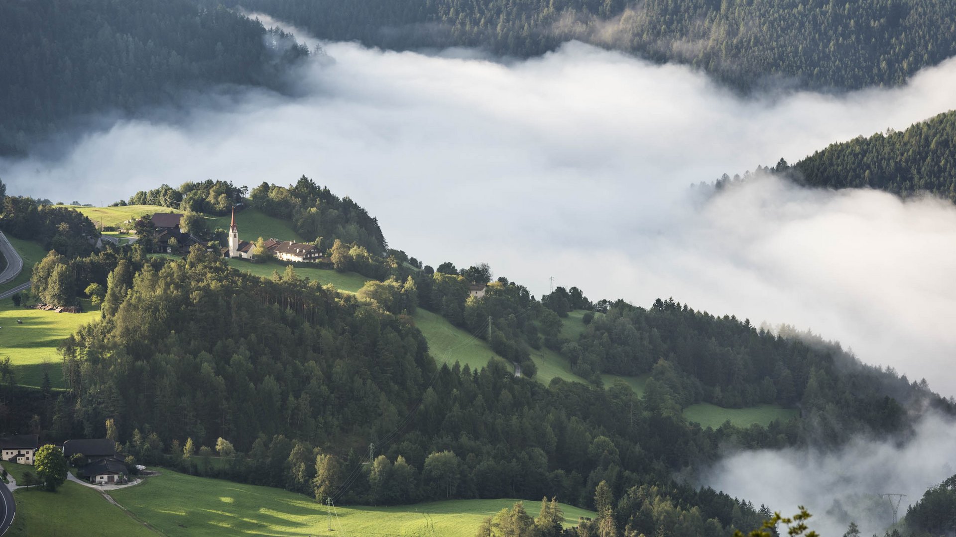 Il paradiso escursionistico di Rodengo L'immagine mostra un paesaggio collinare attraversato da una densa nebbia bianca. Su una delle colline si trova una piccola chiesa, circondata da prati verdi e boschi che emergono dalla nebbia.