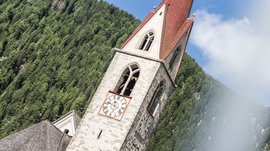 Valles & il villaggio alpino più bello dell’Alto Adige L'immagine mostra un campanile con una punta rossa e due grandi orologi con numeri romani. Sullo sfondo si vedono foreste fitte di conifere verdi e il cielo è parzialmente nuvoloso.