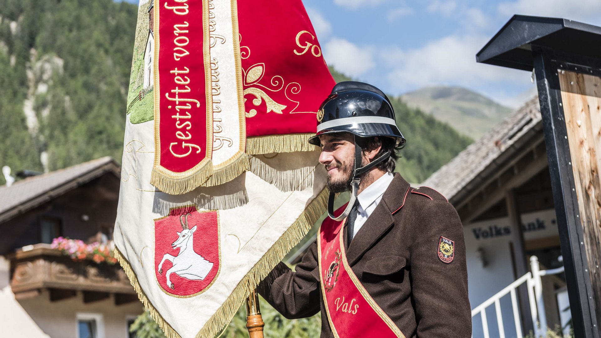 Valles & il villaggio alpino più bello dell’Alto Adige L'immagine mostra un uomo in uniforme marrone con una fascia rossa su cui è scritto "Vals". Indossa un casco nero e tiene una bandiera rosso-bianca con decorazioni dorate e una capra bianca sopra. Sullo sfondo si vedono una casa e delle montagne.