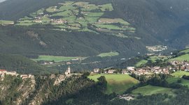 Il paradiso escursionistico di Rodengo L'immagine mostra un paesaggio ampio con un castello e una chiesa che dominano una roccia. Sullo sfondo si estendono campi verdi, foreste e piccoli villaggi in un ambiente collinare.