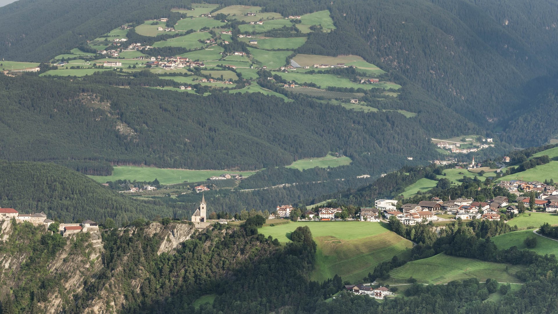 Il paradiso escursionistico di Rodengo L'immagine mostra un paesaggio ampio con un castello e una chiesa che dominano una roccia. Sullo sfondo si estendono campi verdi, foreste e piccoli villaggi in un ambiente collinare.