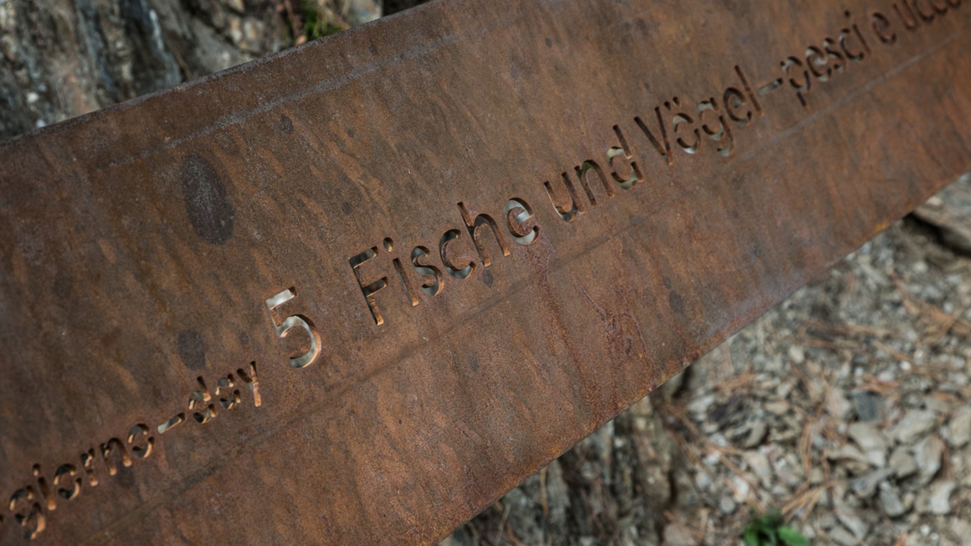 Il Sentiero della Creazione a Rodengo L'immagine mostra una lastra di metallo arrugginita con inciso il testo "Tag 5 pesci e uccelli". Lo sfondo è composto da elementi naturali come terra, pietre e corteccia d'albero.