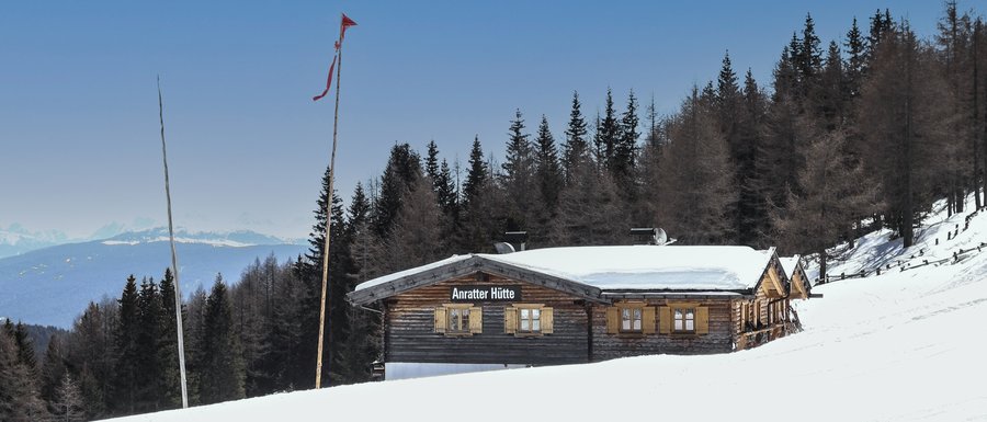 Benvenuti nell’Area Vacanze Sci e Malghe Rio Pusteria! L'immagine mostra la "Anratter Hütte", una baita di legno su un pendio innevato, circondata da conifere. Sullo sfondo si vedono cime innevate sotto un cielo azzurro. Una bandiera rossa sventola su uno dei pennoni davanti alla baita.