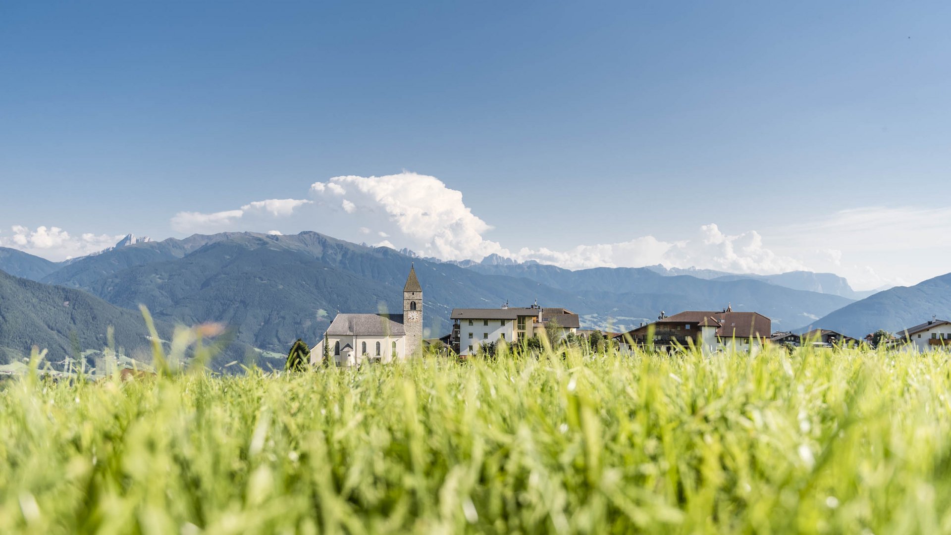 Conoscete già Maranza? L'immagine mostra un prato con erba alta e verde in primo piano. Dietro c'è un piccolo villaggio con una chiesa e diverse case. Sullo sfondo si vedono alte montagne sotto un cielo limpido e azzurro.