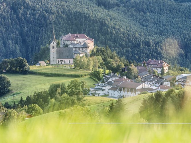 Il paradiso escursionistico di Rodengo L'immagine mostra una chiesa e un edificio su una collina, circondati da prati verdi e foreste dense. In primo piano si vede dell'erba sfocata.