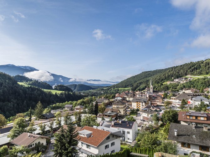 Il paradiso escursionistico di Rodengo L'immagine mostra una pittoresca città circondata da verdi colline e montagne. In primo piano si vedono case con tetti rossi e vegetazione rigogliosa, mentre sullo sfondo si riconoscono montagne avvolte dalla nebbia e un cielo azzurro limpido.