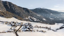 Il paradiso escursionistico di Rodengo L'immagine mostra un piccolo villaggio innevato situato in una paesaggio collinare. Sullo sfondo si ergono montagne boscose, anch'esse coperte da un sottile strato di neve. La scena appare tranquilla e invernale, con un cielo limpido e edifici sparsi che giacciono nella neve.