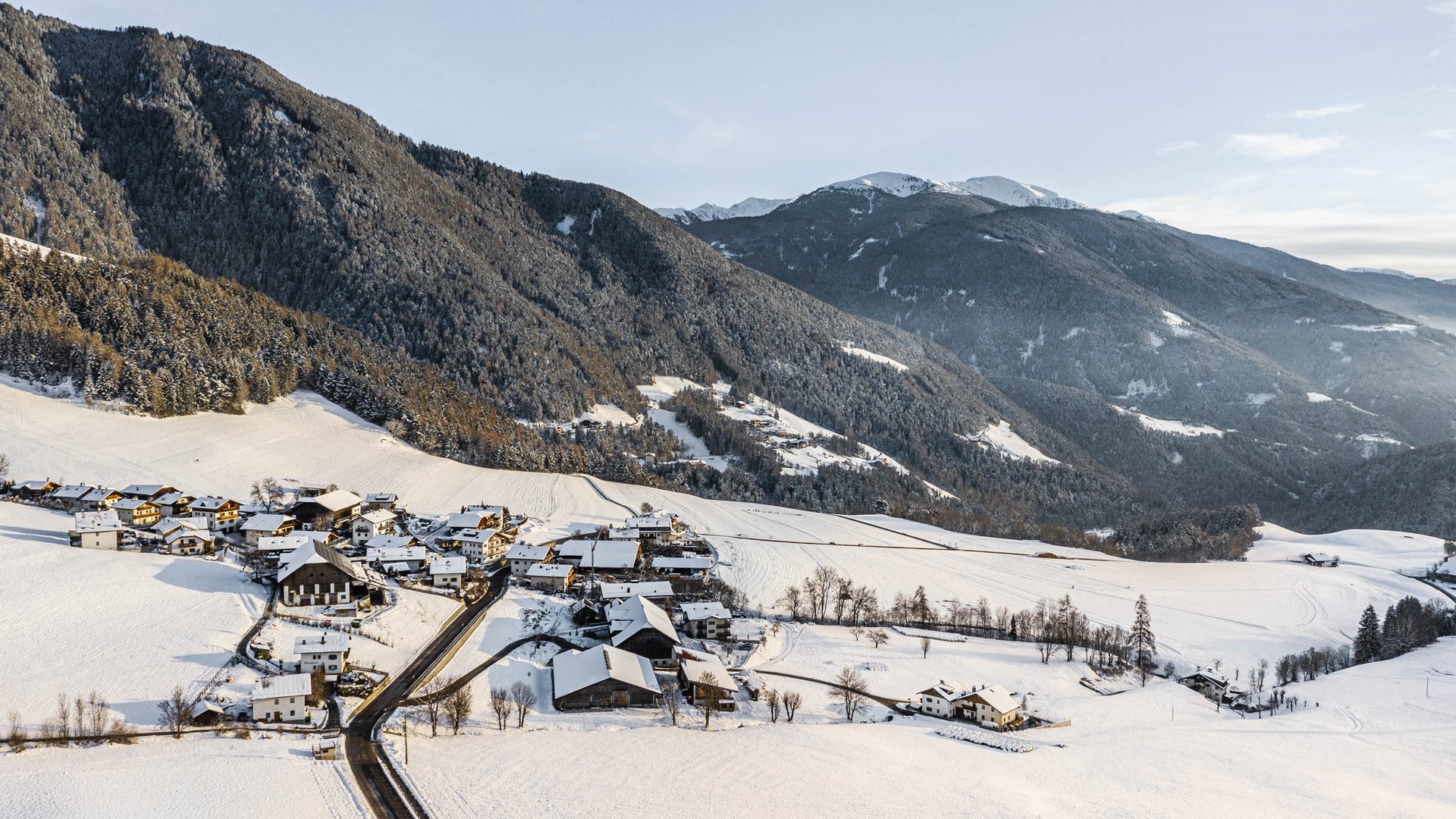 Il paradiso escursionistico di Rodengo L'immagine mostra un piccolo villaggio innevato situato in una paesaggio collinare. Sullo sfondo si ergono montagne boscose, anch'esse coperte da un sottile strato di neve. La scena appare tranquilla e invernale, con un cielo limpido e edifici sparsi che giacciono nella neve.