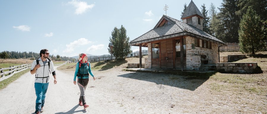 Benvenuti nell’Area Vacanze Sci e Malghe Rio Pusteria! L'immagine mostra due escursionisti, un uomo e una donna, che camminano lungo un ampio sentiero sabbioso. A destra di loro c'è una piccola cappella di pietra con un tetto in legno. Il cielo è limpido e blu, e sullo sfondo si vedono dolci colline e alberi.
