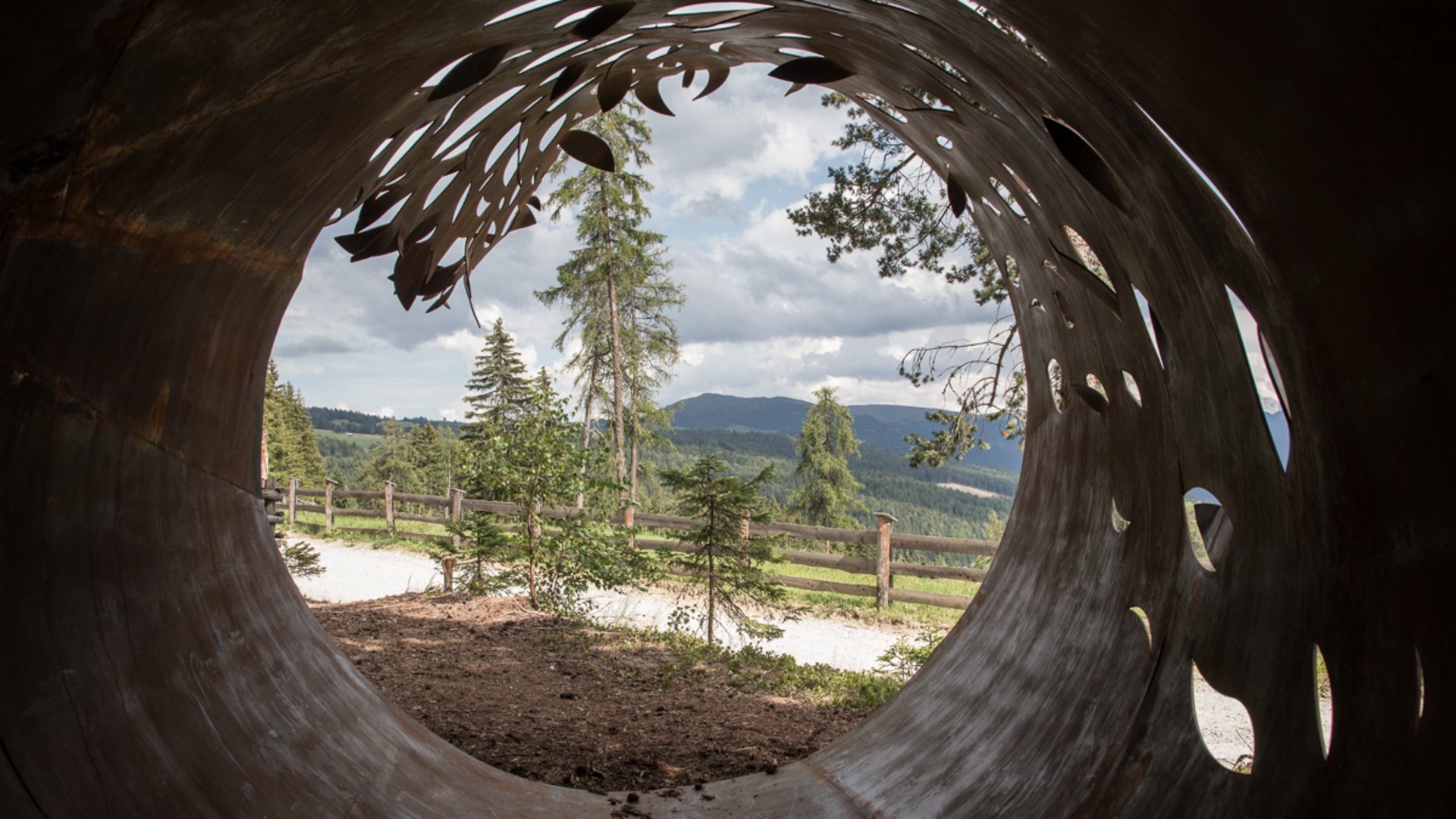 Il Sentiero della Creazione a Rodengo L'immagine mostra la vista attraverso un grande tubo metallico color ruggine, artisticamente traforato con motivi di foglie. Fuori si vedono alberi, una recinzione in legno e in lontananza colline sotto un cielo coperto di nuvole.