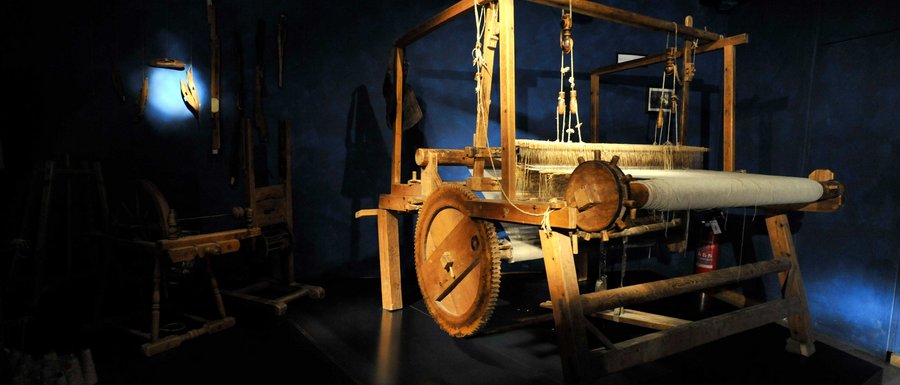 Benvenuti nell’Area Vacanze Sci e Malghe Rio Pusteria! L'immagine mostra un grande telaio in legno in una stanza buia, illuminata da un raggio di luce. Sullo sfondo si riconoscono altri utensili storici appesi al muro.