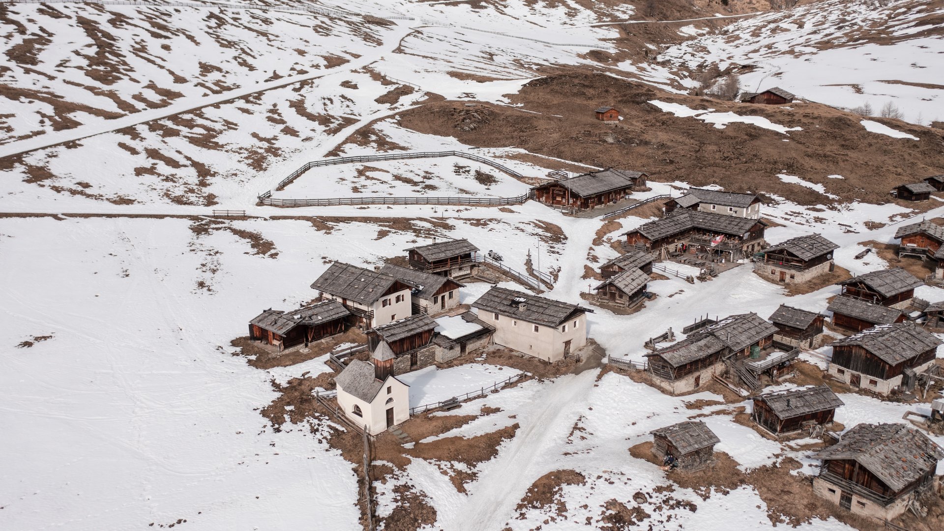 Malga Fane, il più bel villaggio alpino dell’Alto Adige L'immagine mostra un piccolo villaggio in un paesaggio montano innevato. Le case hanno per lo più tetti in legno o pietra, e le colline circostanti sono in parte coperte di neve, mentre si vedono delle aree marroni.