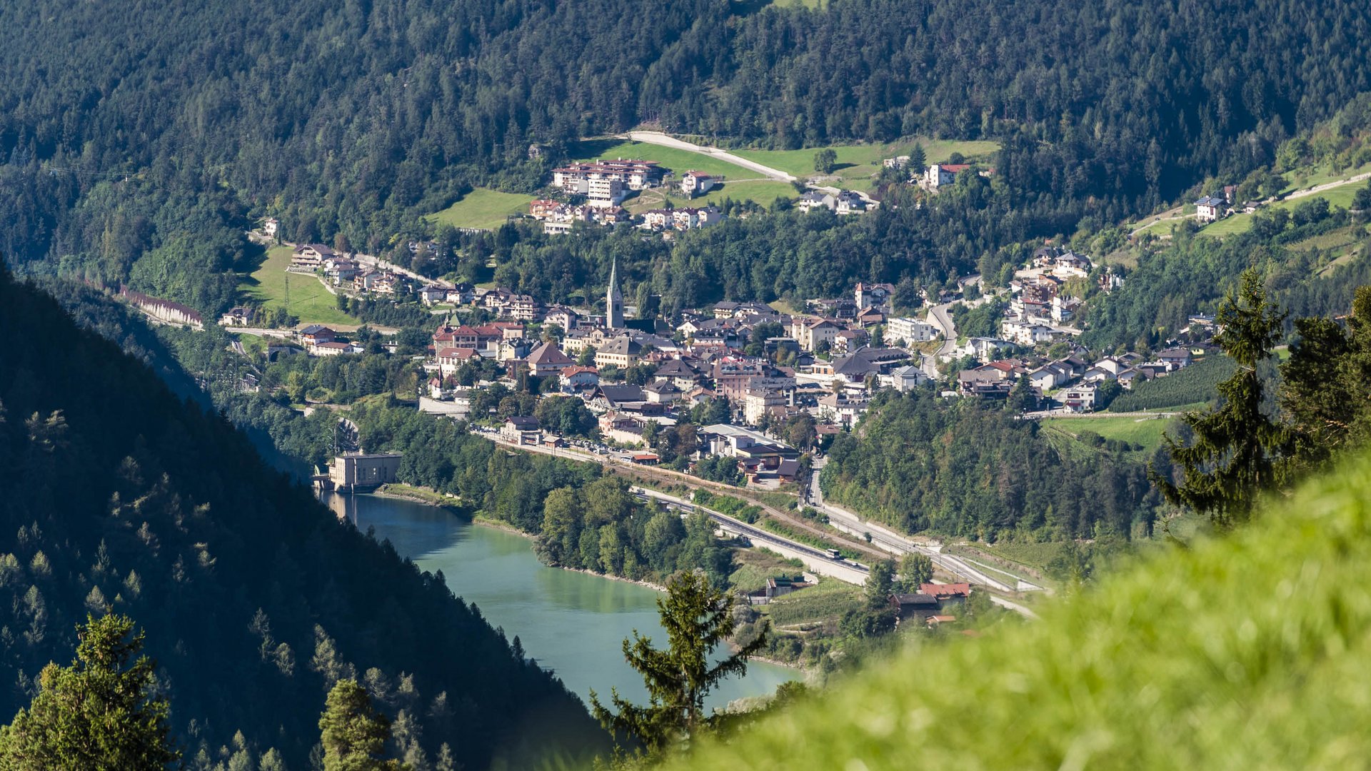 Alla scoperta di Rio di Pusteria L'immagine mostra una piccola città che si estende in una valle verde, circondata da colline densamente boscose. Nel centro della città spicca un campanile, e in primo piano è visibile un lago, sulle cui rive si trova un edificio.