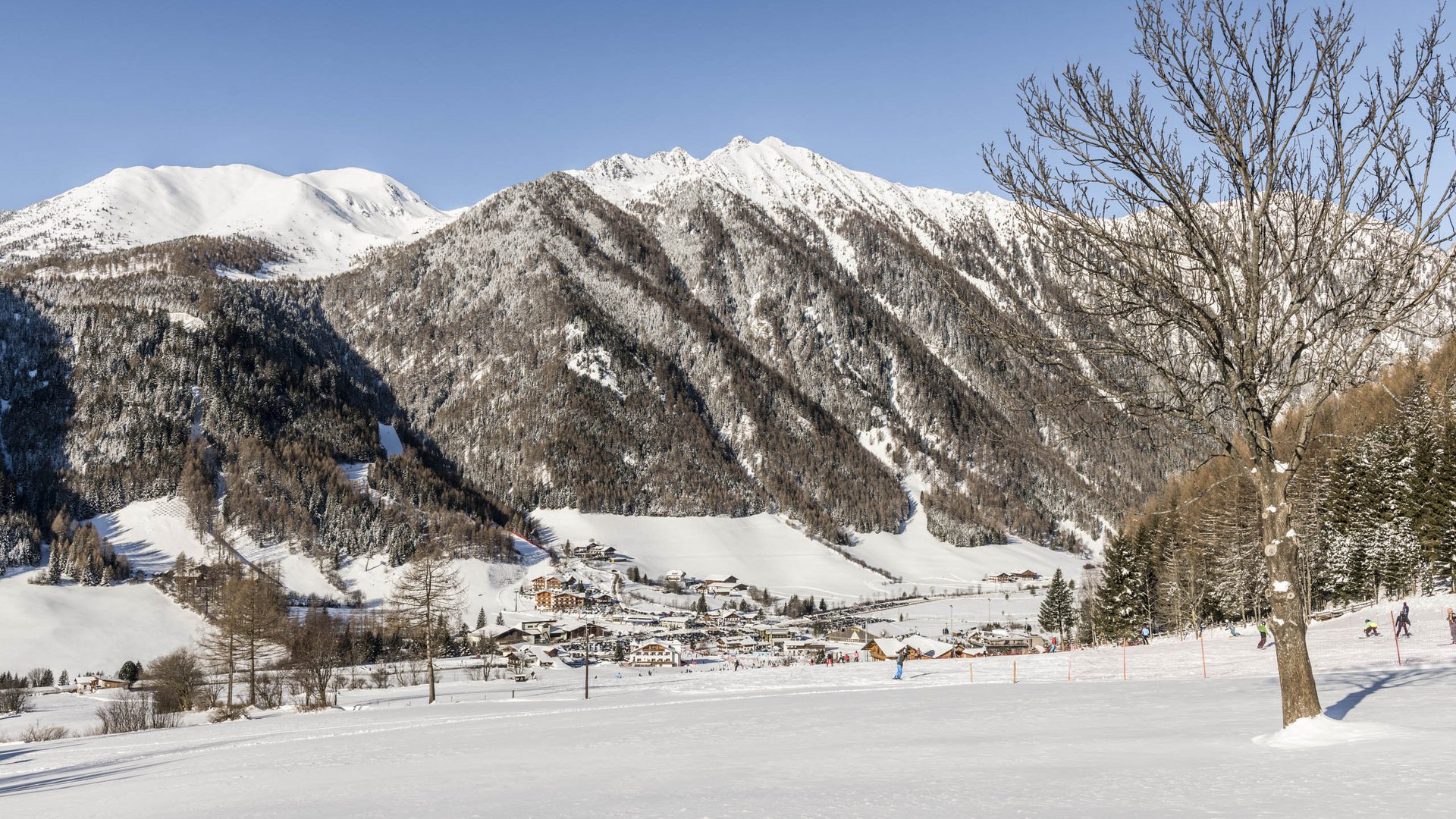 Valles & il villaggio alpino più bello dell’Alto Adige L'immagine mostra un paesaggio montano invernale con cime innevate sullo sfondo e un piccolo villaggio nella valle. In primo piano si vede un albero spoglio, mentre persone passeggiano o sciano nella neve.