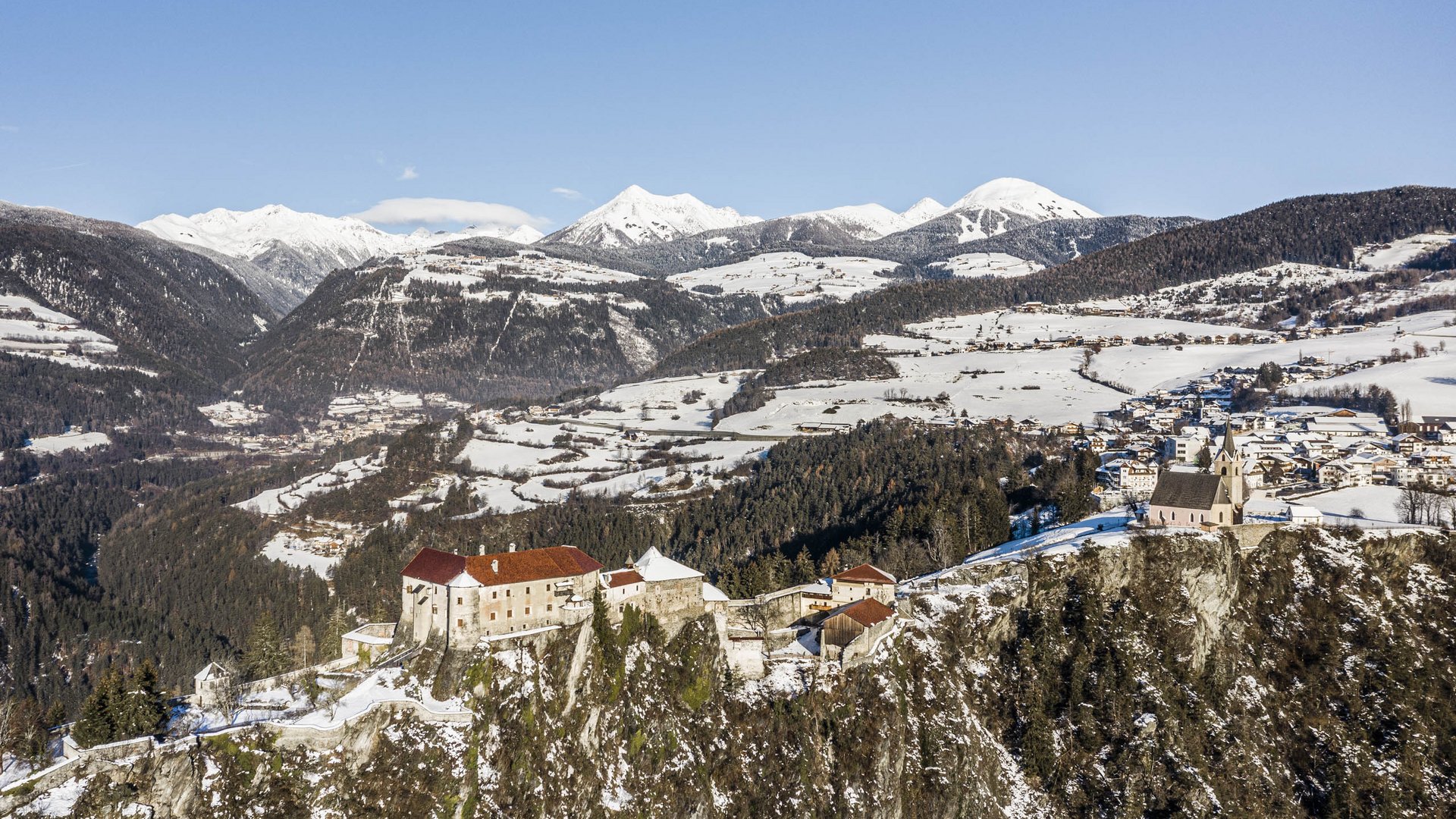 Il paradiso escursionistico di Rodengo L'immagine mostra un castello che domina una roccia scoscesa e coperta di neve, circondato da un paesaggio montano invernale. Sullo sfondo si estendono montagne innevate sotto un cielo blu chiaro, mentre in valle si vedono villaggi sparsi e foreste.