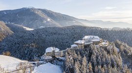 Il paradiso escursionistico di Rodengo L'immagine mostra un castello situato su una collina boscosa coperta di neve. Il castello stesso ha tetti innevati ed è circondato da foreste fitte. Sullo sfondo si estendono dolci colline e montagne sotto un cielo limpido e azzurro.