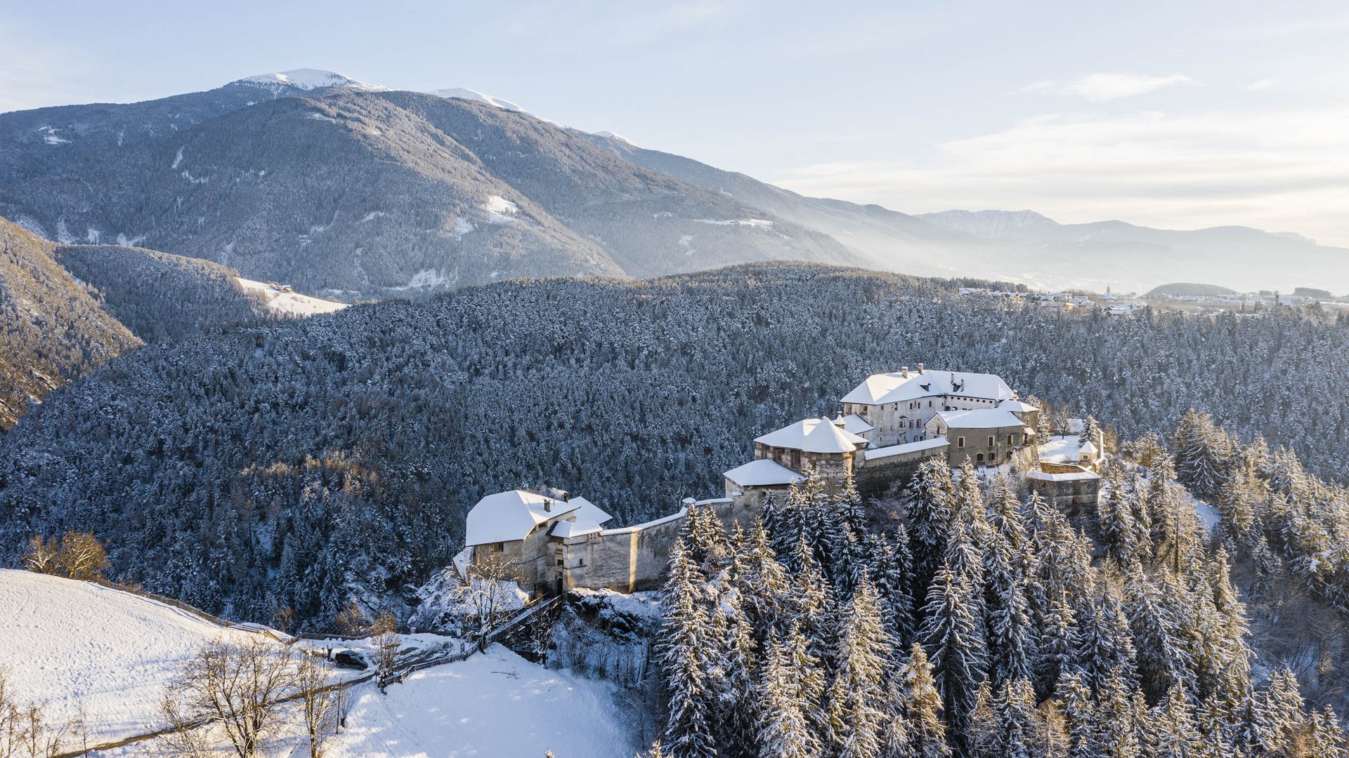 Il paradiso escursionistico di Rodengo L'immagine mostra un castello situato su una collina boscosa coperta di neve. Il castello stesso ha tetti innevati ed è circondato da foreste fitte. Sullo sfondo si estendono dolci colline e montagne sotto un cielo limpido e azzurro.