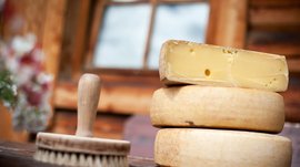 Malga Fane, il più bel villaggio alpino dell’Alto Adige L'immagine mostra diverse forme di formaggio, di cui una tagliata, permettendo di vedere l'interno giallo con alcuni buchi. Accanto c'è una spazzola rotonda su un tavolo di legno, sullo sfondo si intravede sfocato una finestra.