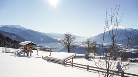 Il paradiso escursionistico di Rodengo L'immagine mostra un paesaggio innevato con una recinzione di legno, una piccola casetta di legno e alberi in primo piano. Sullo sfondo si estende una valle ampia, circondata da alte montagne innevate, mentre il sole splende luminoso nel cielo azzurro.