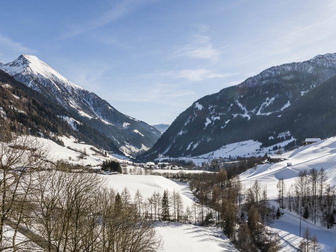 Il paradiso escursionistico di Rodengo L'immagine mostra un paesaggio montano invernale con pendii coperti di neve e una valle al centro. Il sole splende, il cielo è limpido e azzurro, e in primo piano si vedono alberi spogli. Le montagne sullo sfondo sono anch'esse coperte di neve, il che trasmette un'atmosfera di pace e tranquillità.