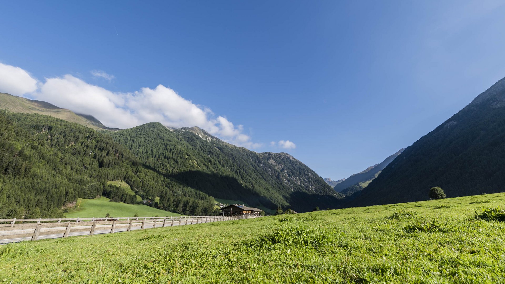 Valles & il villaggio alpino più bello dell’Alto Adige L'immagine mostra un'ampia prateria verde in primo piano, delimitata da una recinzione in legno. Sullo sfondo si ergono pendii montuosi coperti di boschi sotto un cielo azzurro limpido con qualche nuvola.