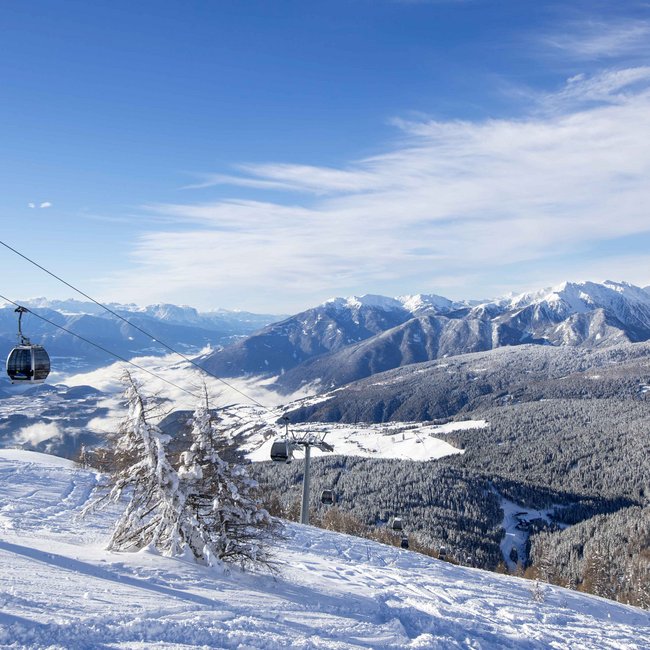 Il comprensorio sciistico Rio Pusteria vi aspetta! Cabine della funivia sopra montagne innevate e alberi sotto un cielo blu