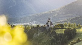 Il paradiso escursionistico di Rodengo L'immagine mostra una chiesa su una roccia, circondata da alberi verdi e prati, con montagne sullo sfondo. Il sole diffonde una luce soffusa sulla scena, rendendo il paesaggio pacifico e idilliaco.