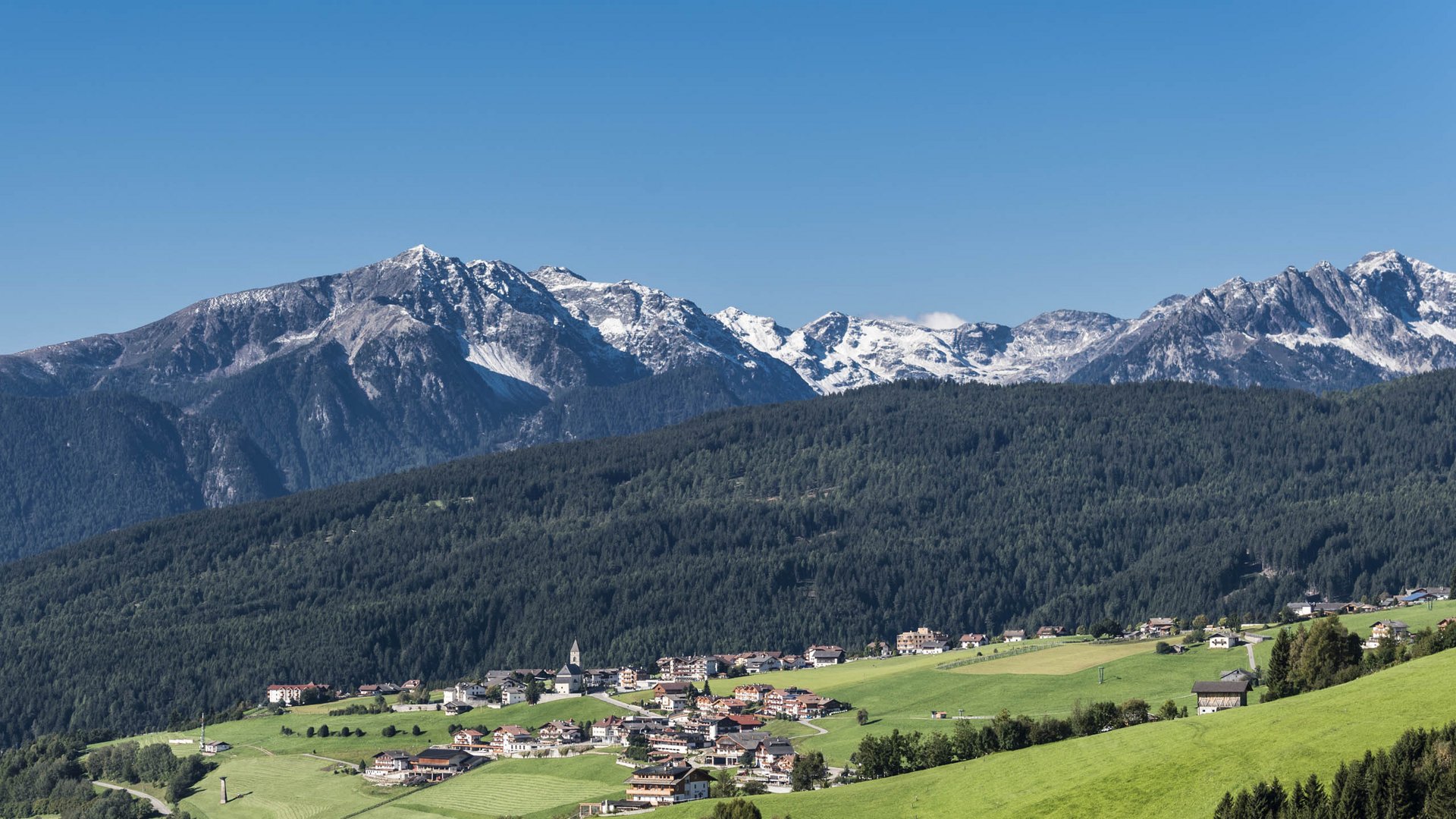Conoscete già Maranza? L'immagine mostra un villaggio che si estende su dolci colline verdi, con sullo sfondo fitte foreste e montagne innevate. Al centro del villaggio si trova una chiesa con una torre appuntita, circondata da case, e l'intera scena è sormontata da un cielo azzurro limpido.