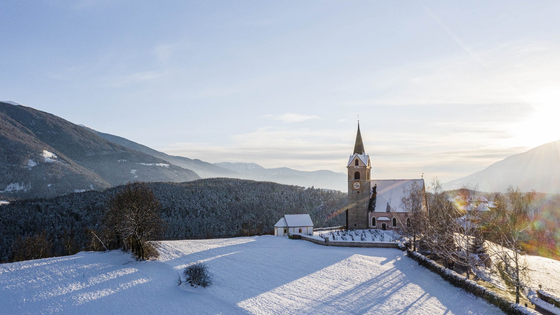 Il paradiso escursionistico di Rodengo L'immagine mostra una chiesa con una torre alta, situata in un paesaggio innevato. Sullo sfondo si vedono colline e montagne boschive sotto un cielo limpido, mentre il sole è basso all'orizzonte e proietta lunghe ombre.