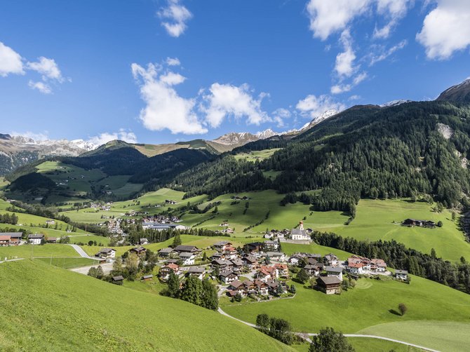 Il paradiso escursionistico di Rodengo L'immagine mostra un paesaggio alpino pittoresco con un piccolo villaggio circondato da prati verdi e colline boschive. Sullo sfondo si vedono montagne innevate sotto un cielo blu limpido con alcune nuvole.