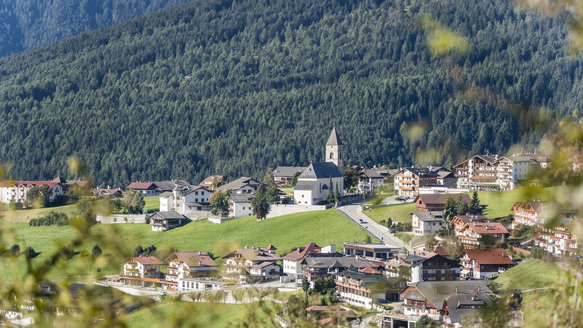 Conoscete già Maranza? L'immagine mostra un villaggio idilliaco con una chiesa centrale e una torre a punta, circondato da case residenziali e edifici. Il villaggio si trova in una valle verde, circondata da pendii montuosi fitti e boscosi. La vegetazione in primo piano è leggermente sfocata, il che conferisce profondità all'immagine.