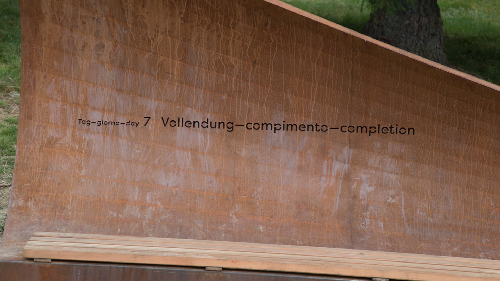 Il Sentiero della Creazione a Rodengo L'immagine mostra una panchina o scultura metallica, sulla quale è inciso il testo "Tag 7 – Vollendung" in diverse lingue. La struttura ha un colore ruggine ed è collocata all'aperto, probabilmente in un ambiente naturale preservato.
